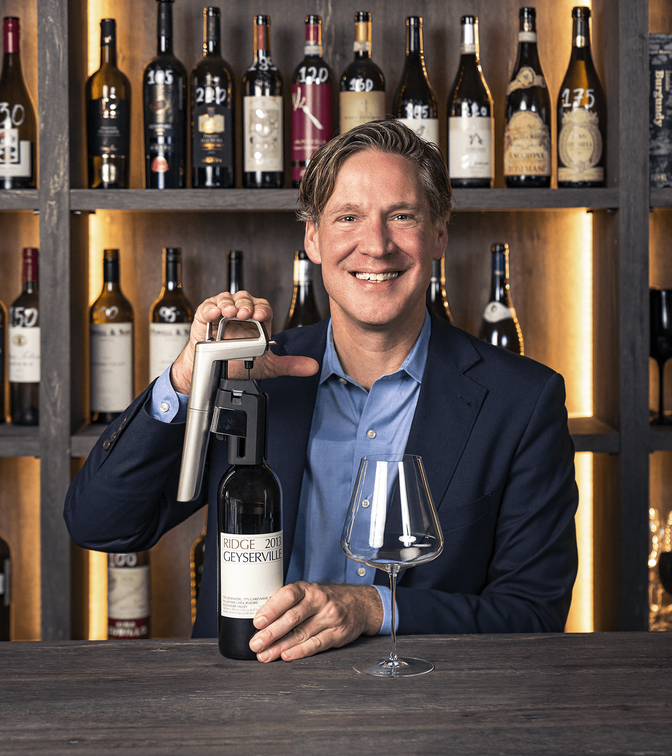 Smiling man holding a wine bottle labeled "Ridge 2013 Geyserville" and a wine opener, with glasses and wine bottles in background.