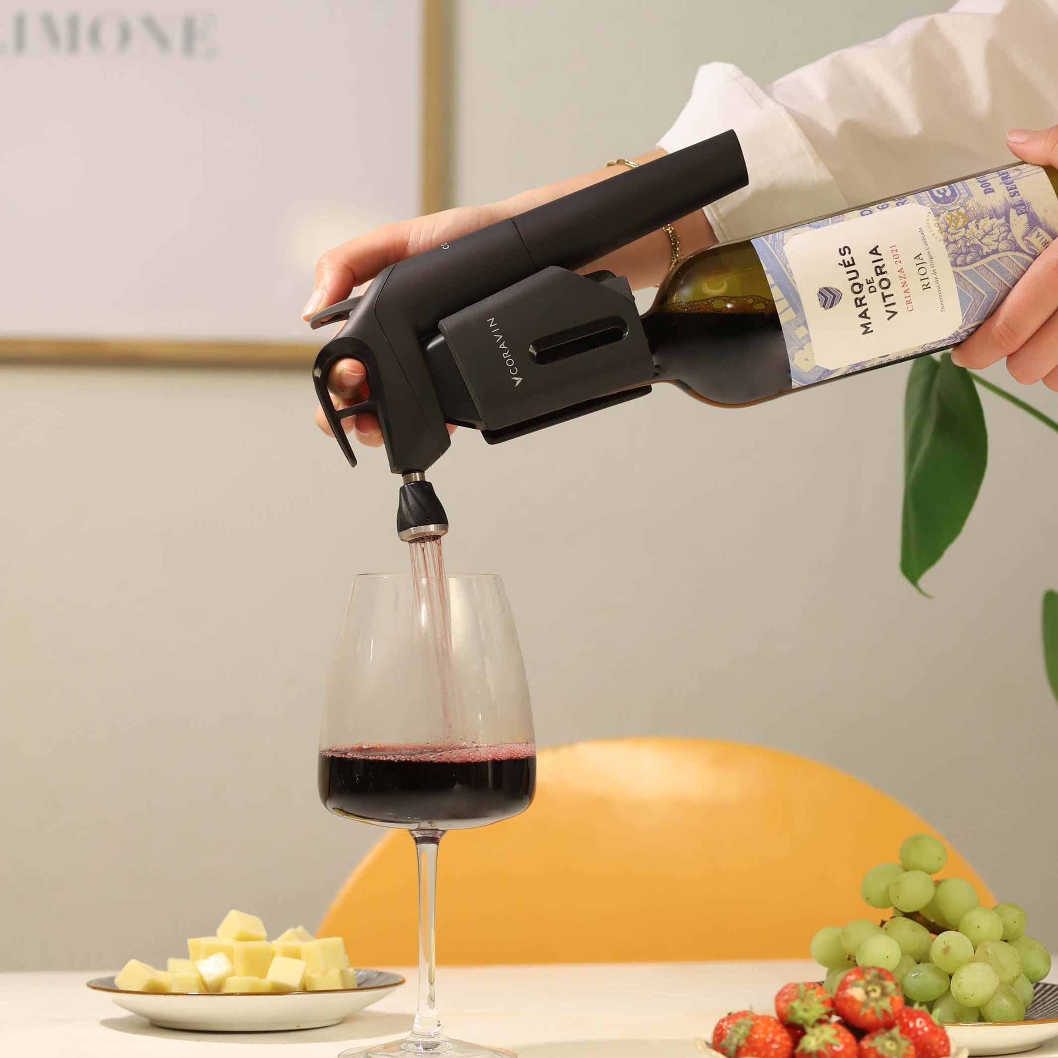 Close-up of a person using a Coravin wine preservation system to pour red wine from a bottle labeled 'Marqués de Vitoria' into a glass, with plates of cheese cubes, green grapes, and strawberries on a table in the foreground.