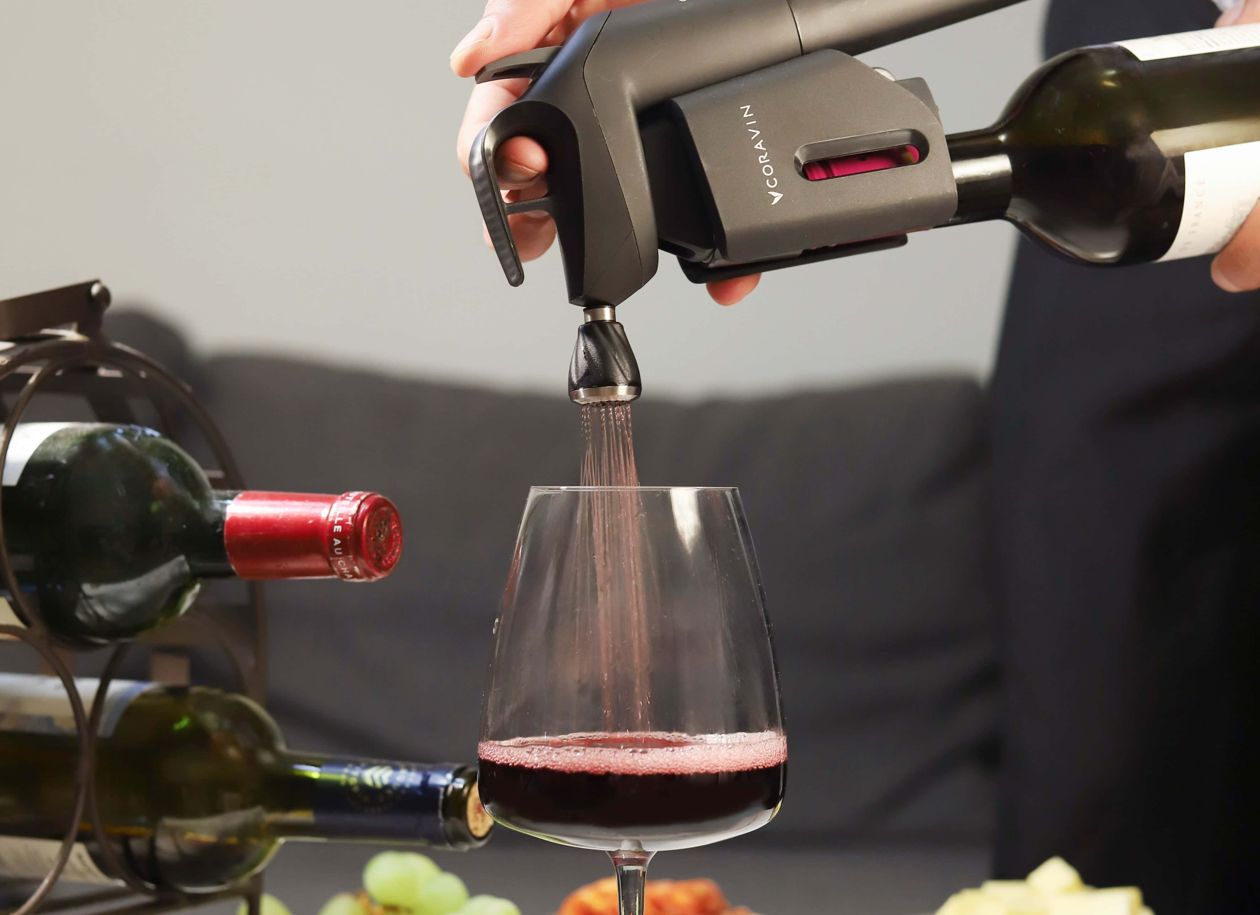 Close-up of Coravin wine preservation system pouring red wine into a glass, with a wine rack and grapes in the background.