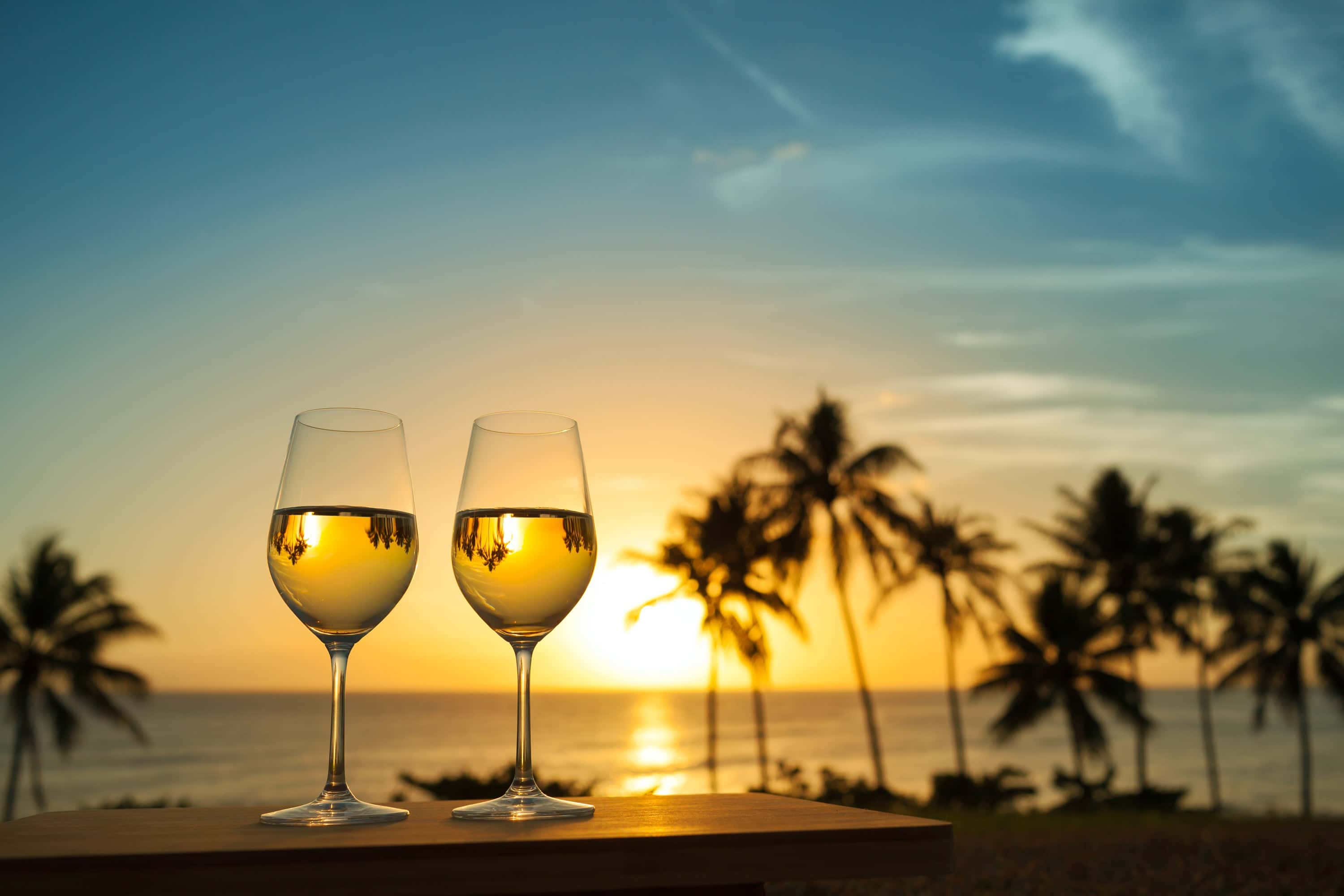 Two glasses of white wine at sunset with a tropical beach and palm trees in the background, evoking a romantic and serene summer atmosphere.