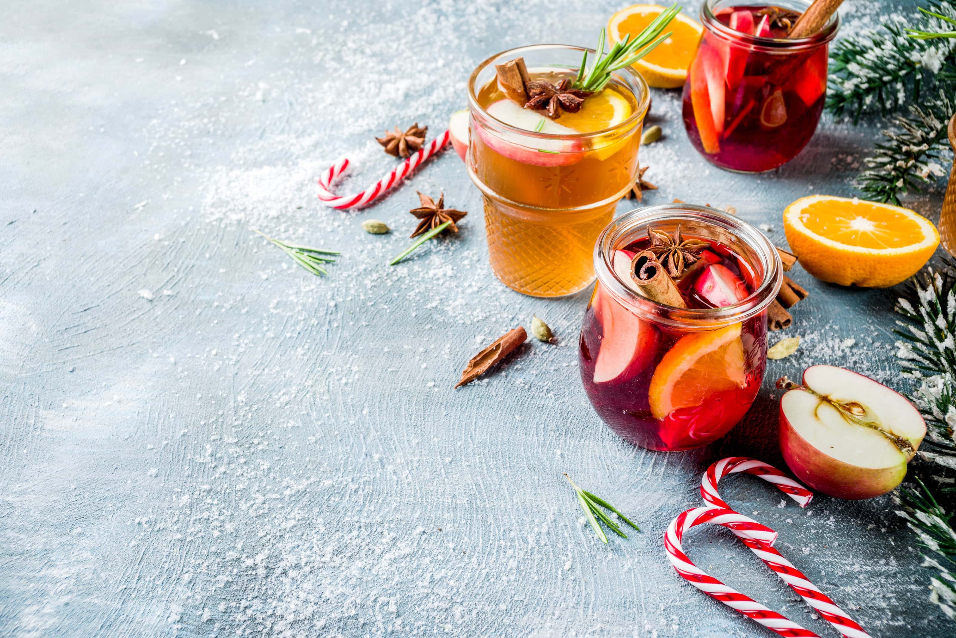 Assorted glasses of holiday mulled wine and cider garnished with fruit slices, cinnamon sticks, and star anise, surrounded by candy canes, orange halves, and snowy evergreen branches.