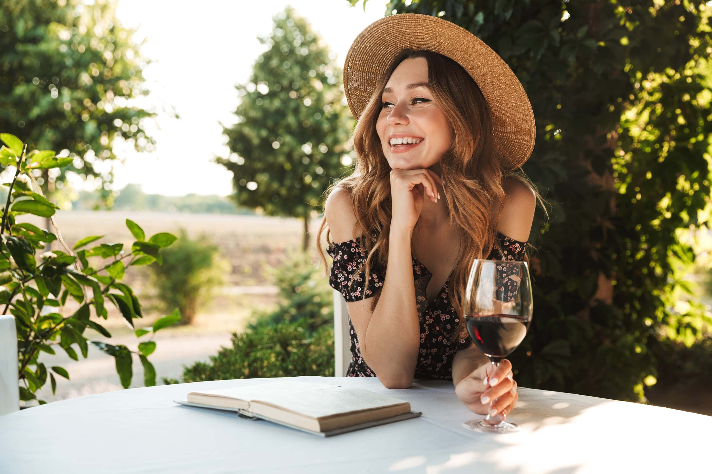 Smiling woman in a summer dress and straw hat enjoying a glass of red wine outdoors with a journal on the table, surrounded by greenery and vineyard views.
