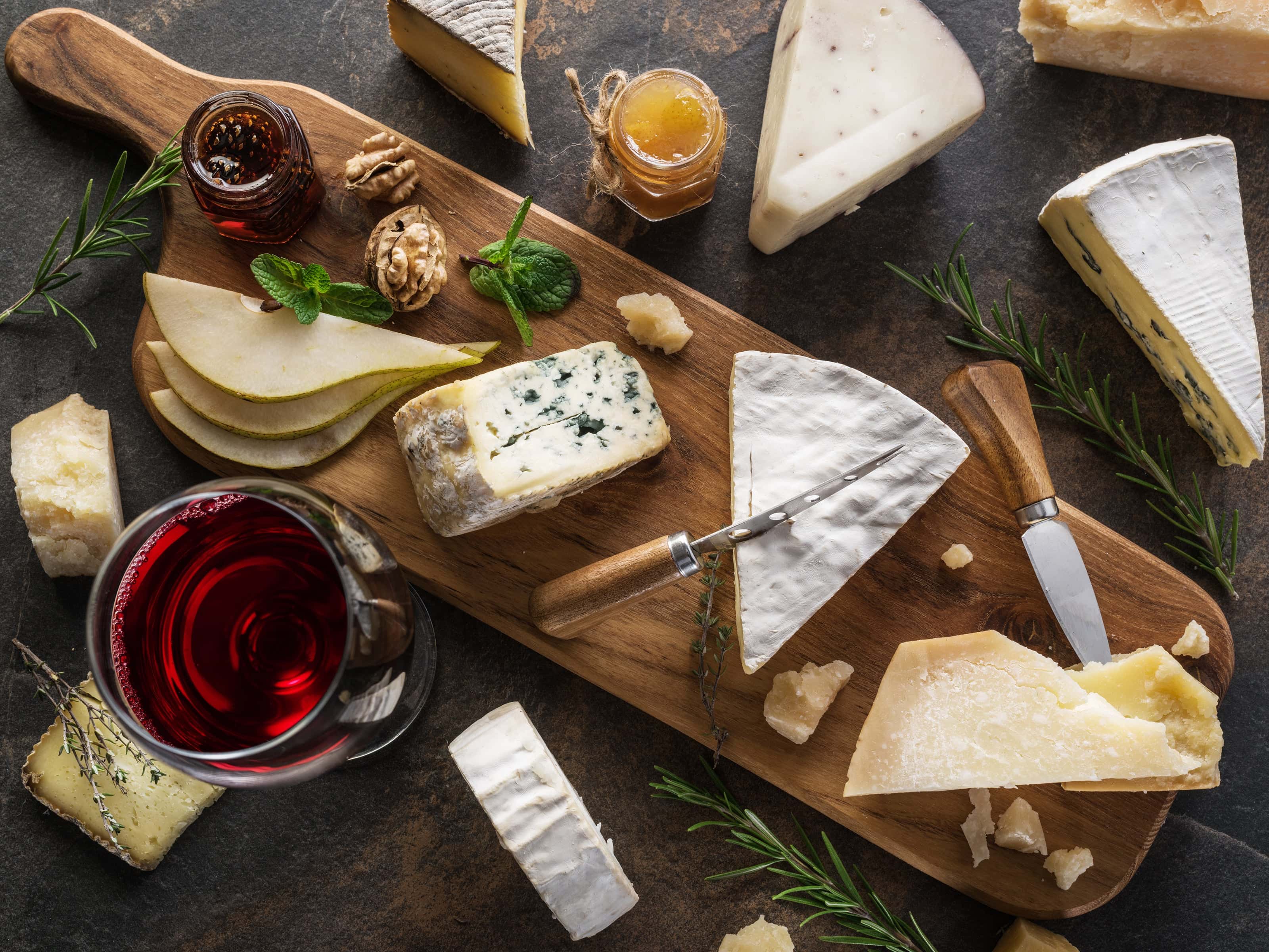 Assorted gourmet cheeses with pear slices, walnuts, fig jam, and a glass of red wine on a rustic wooden serving board.