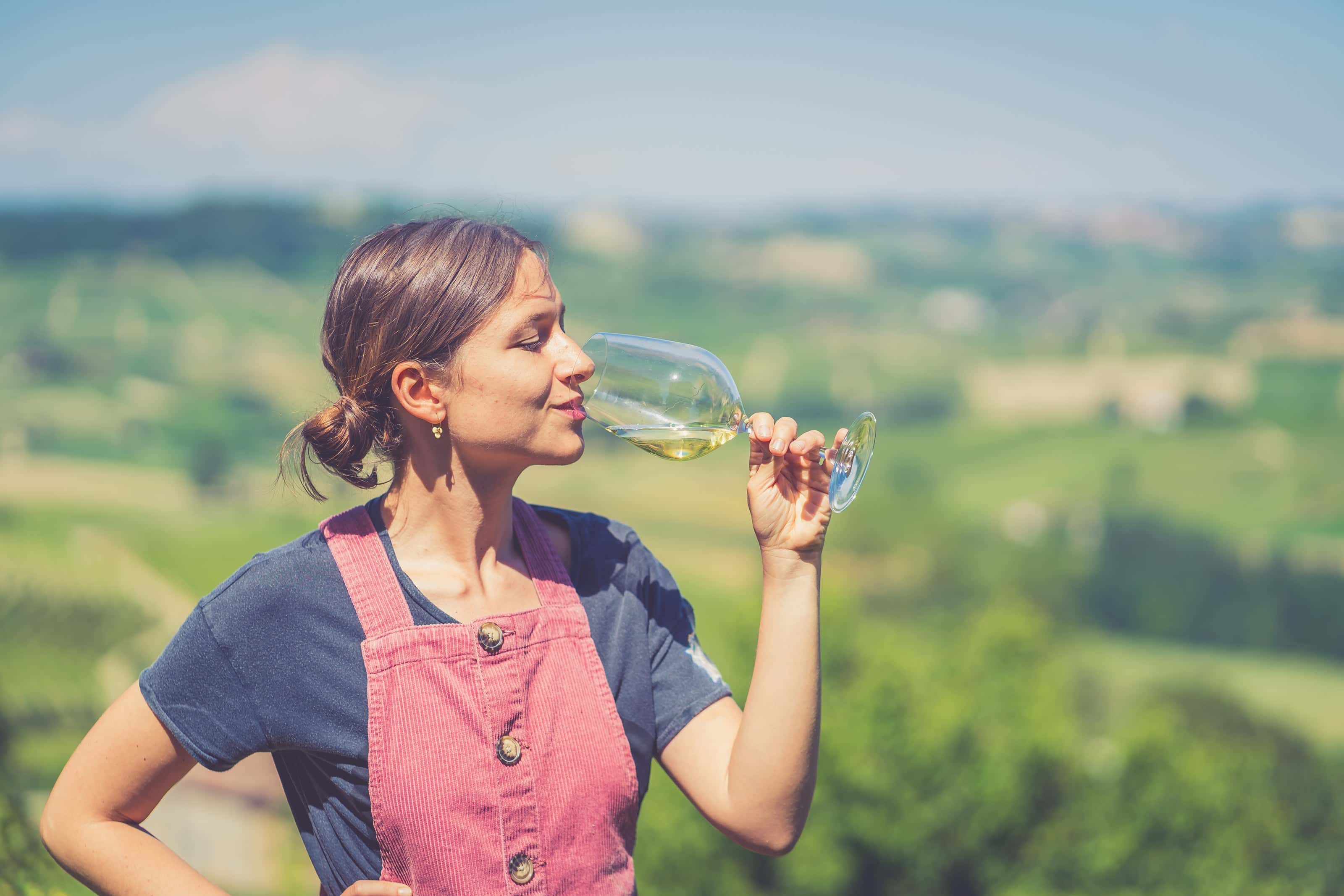 Young woman enjoying a glass of white wine outdoors with vineyard landscape in the background on a sunny day.