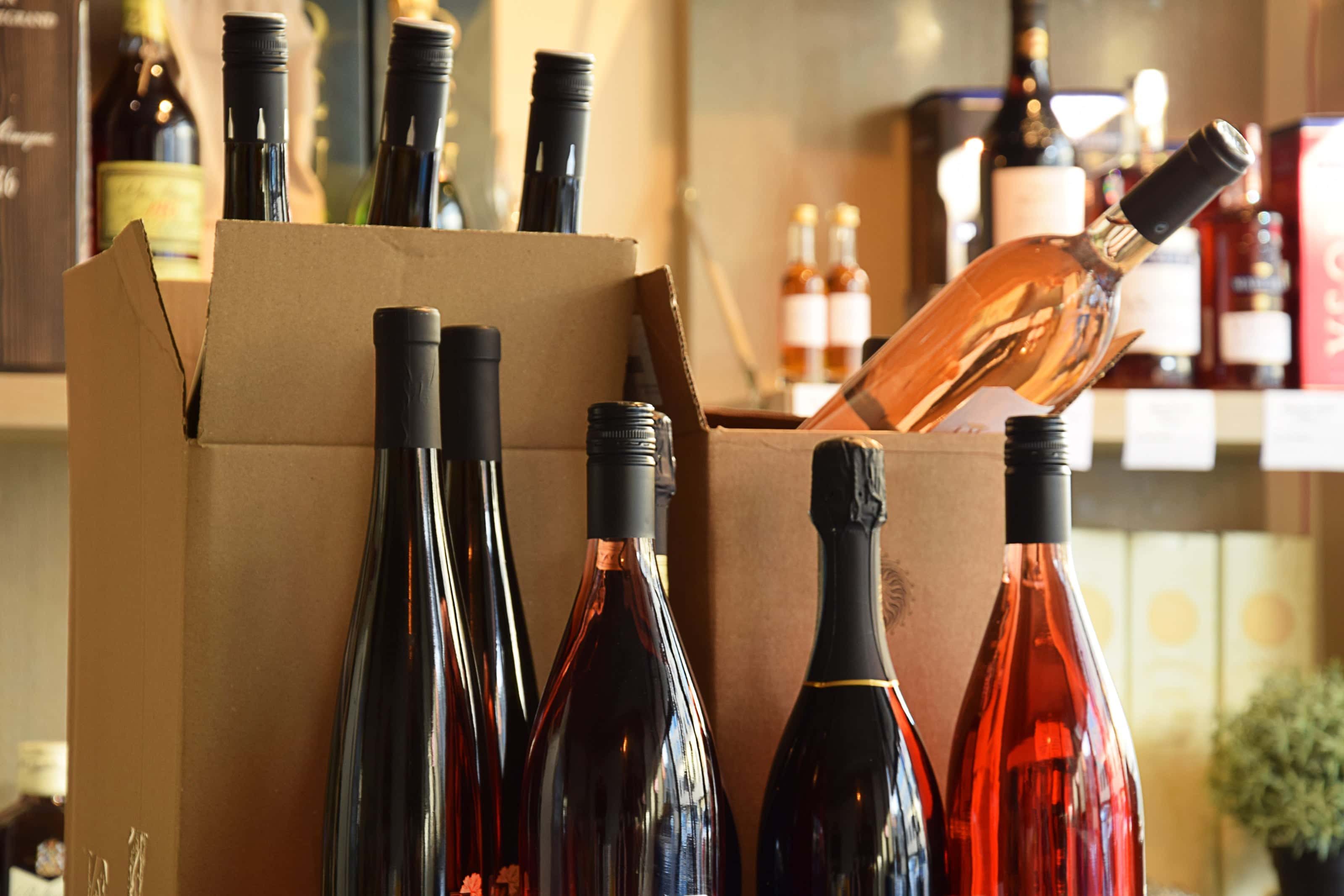 Cardboard boxes filled with assorted wine bottles, including rosé and red varieties, set on a counter in a wine shop with shelves of additional bottles in the background.