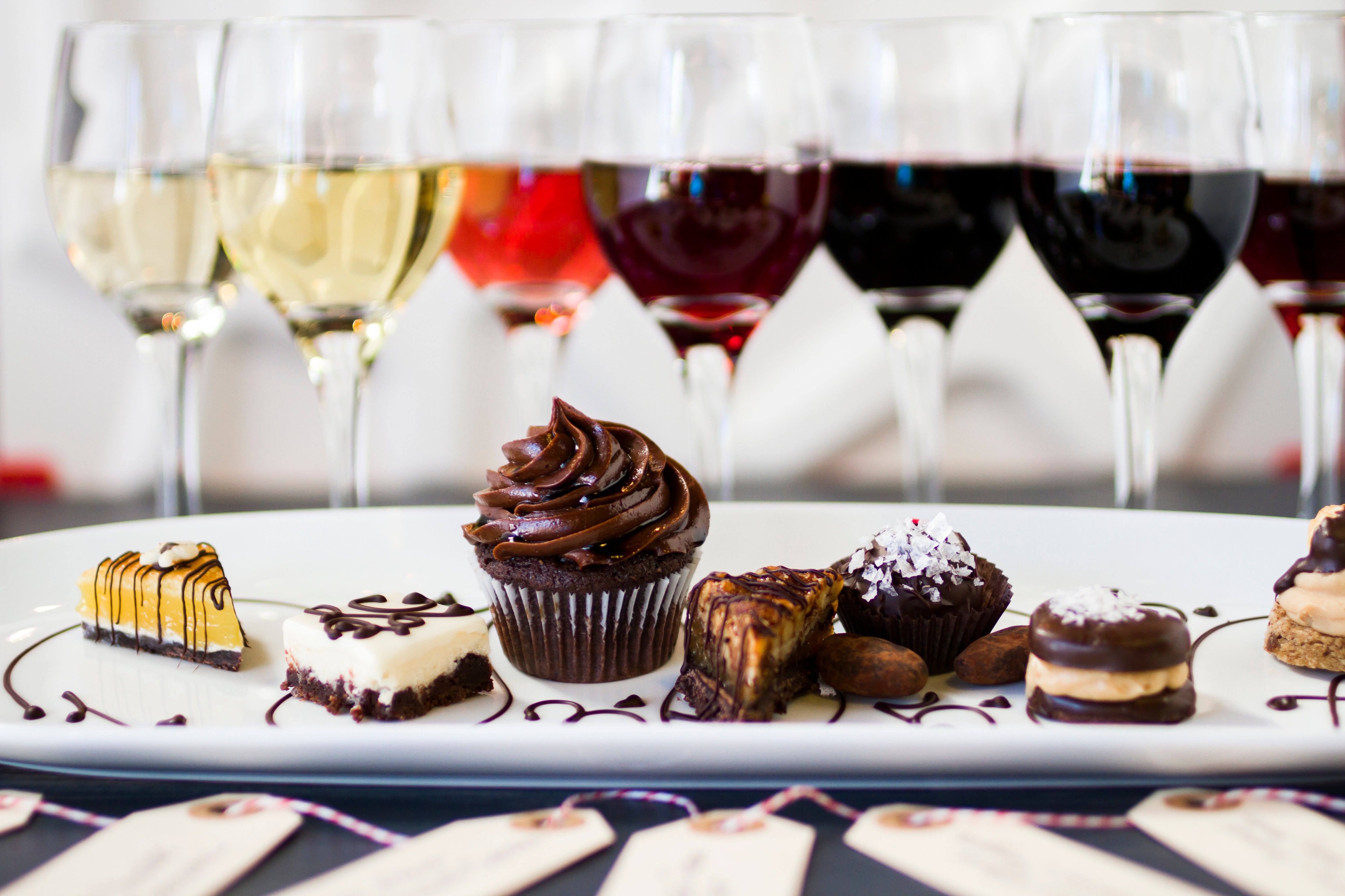 A dessert and wine pairing display featuring assorted chocolate treats and pastries on a white plate, with glasses of white, rosé, and red wine lined up in the background.