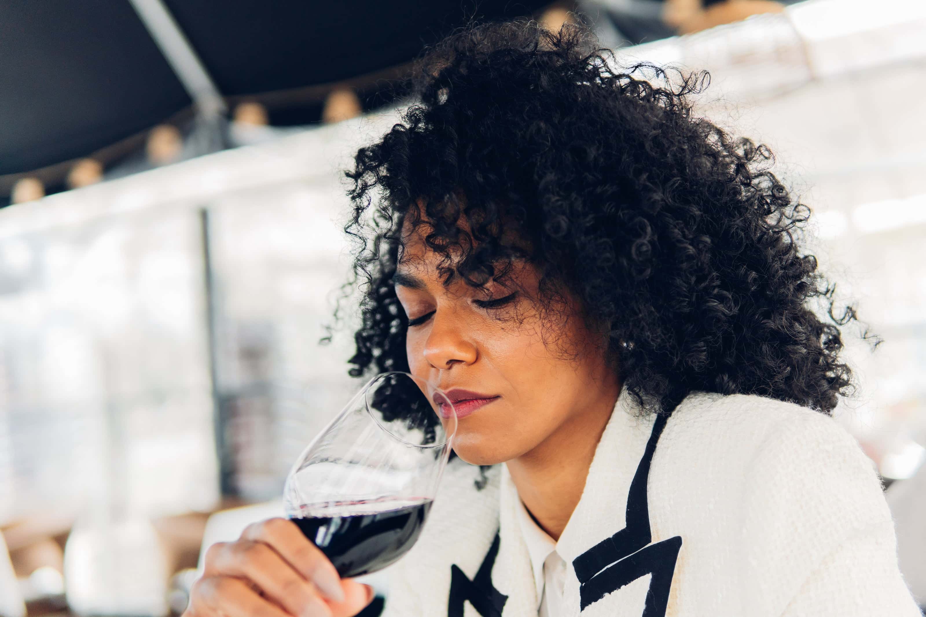 Woman with curly hair and a white jacket thoughtfully smelling a glass of red wine at a modern, well-lit indoor setting.