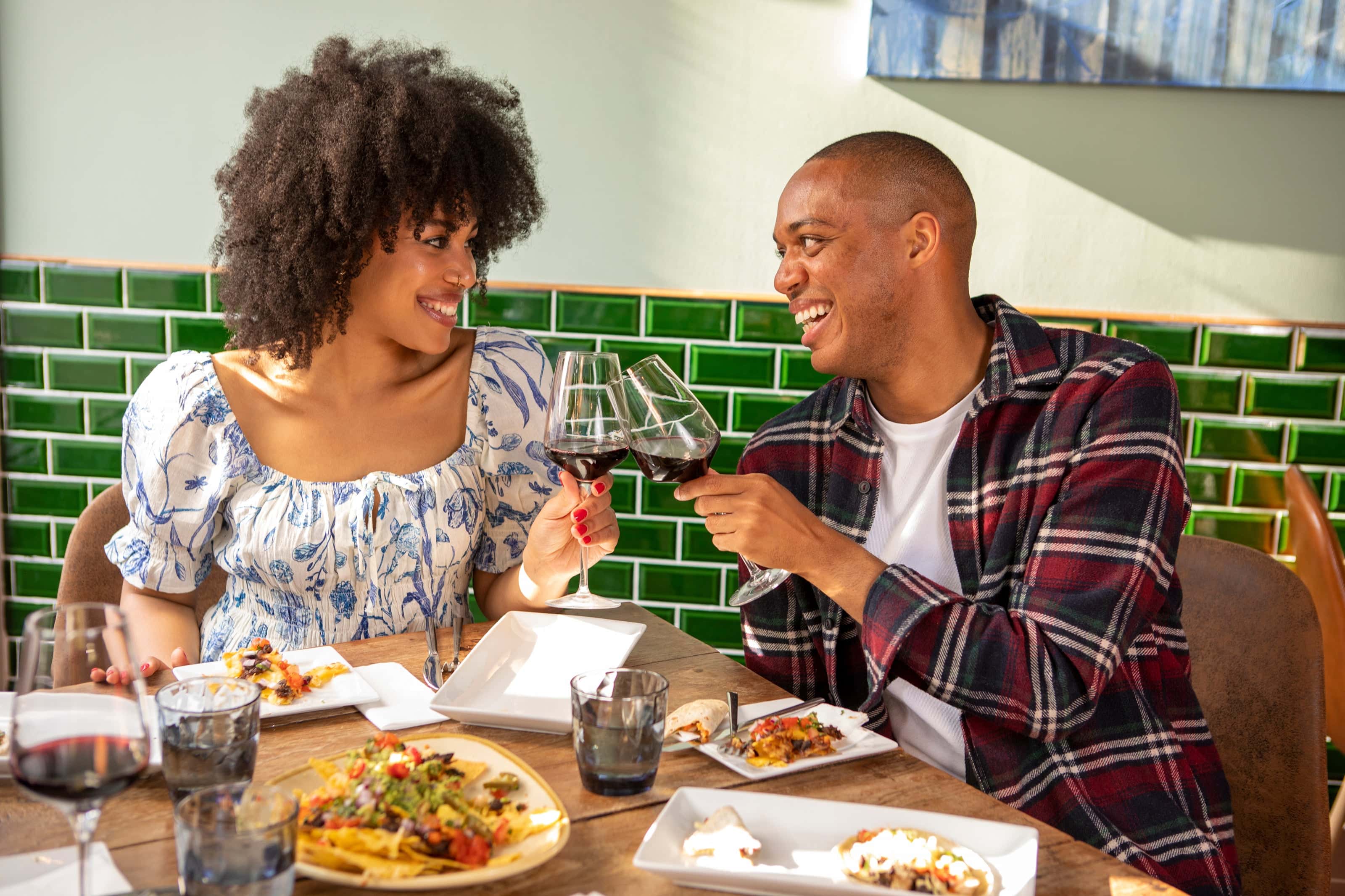 Smiling couple toasting glasses of red wine over a vibrant lunch spread at a sunlit restaurant with green tiled walls.