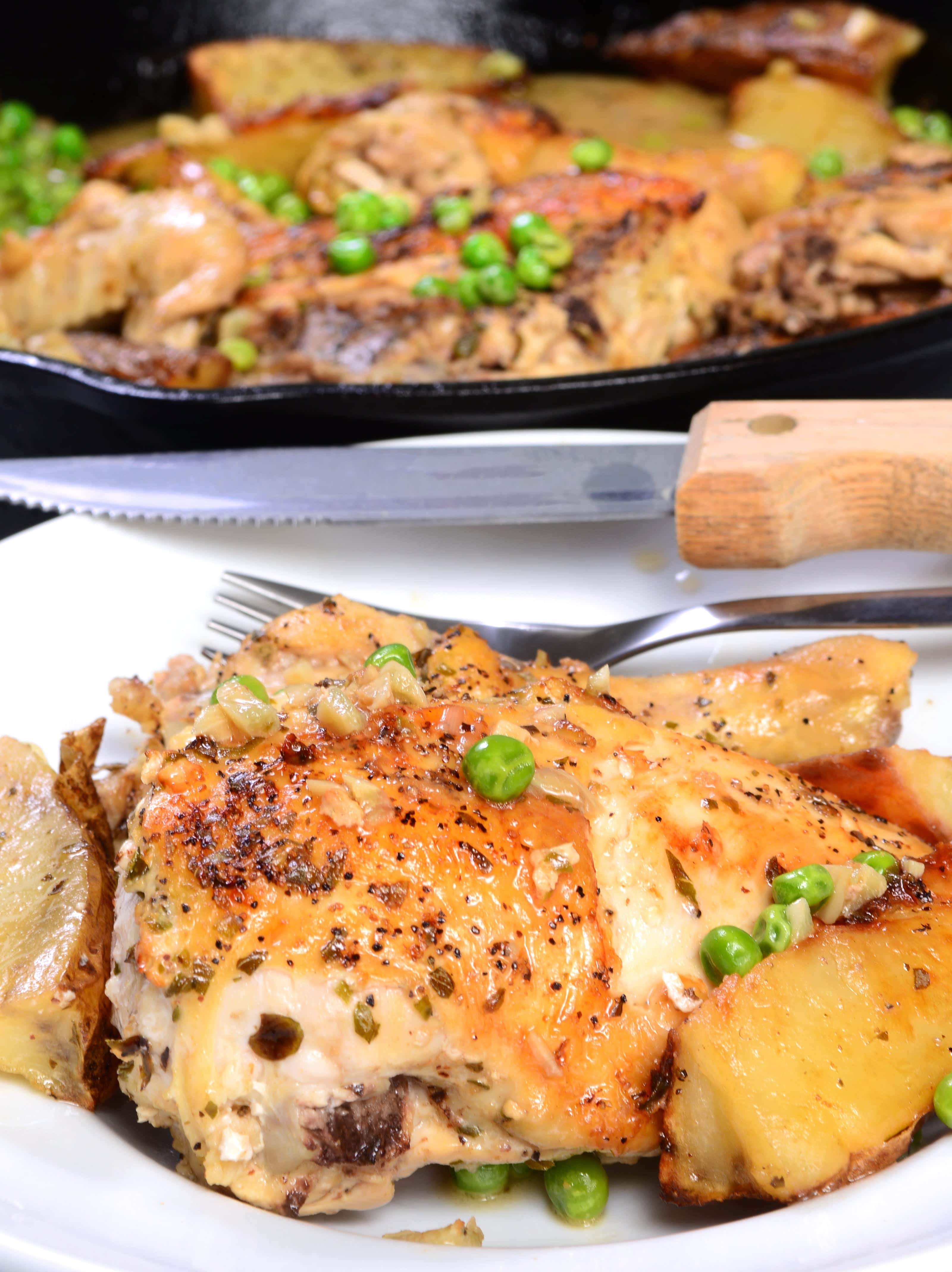 Close-up of a roasted chicken thigh served with golden-brown potatoes and green peas on a white plate, with a cast iron skillet filled with more of the dish in the background.