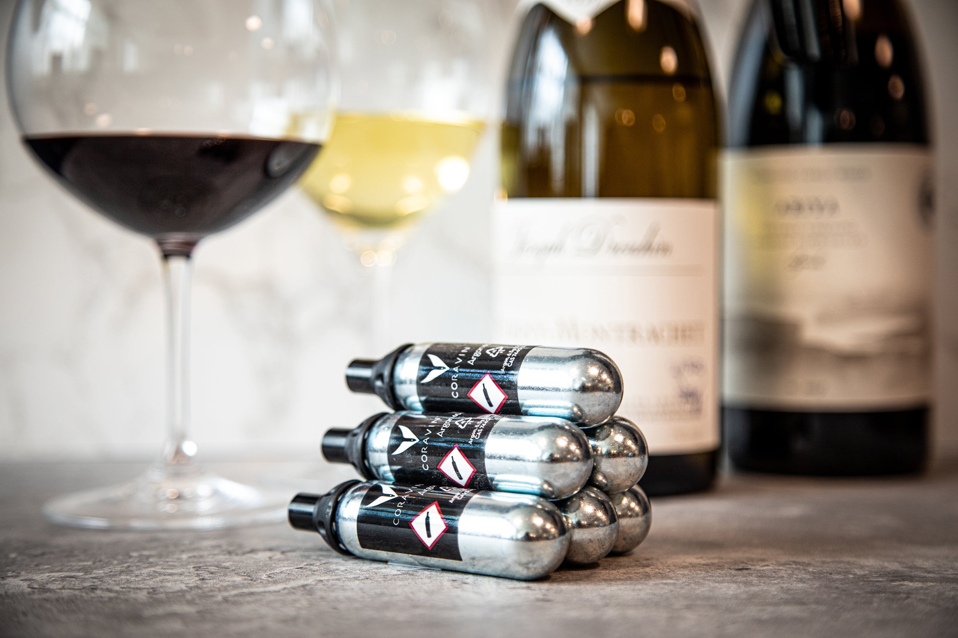 Stack of Coravin argon gas capsules on a countertop with glasses of red and white wine and wine bottles in the background.