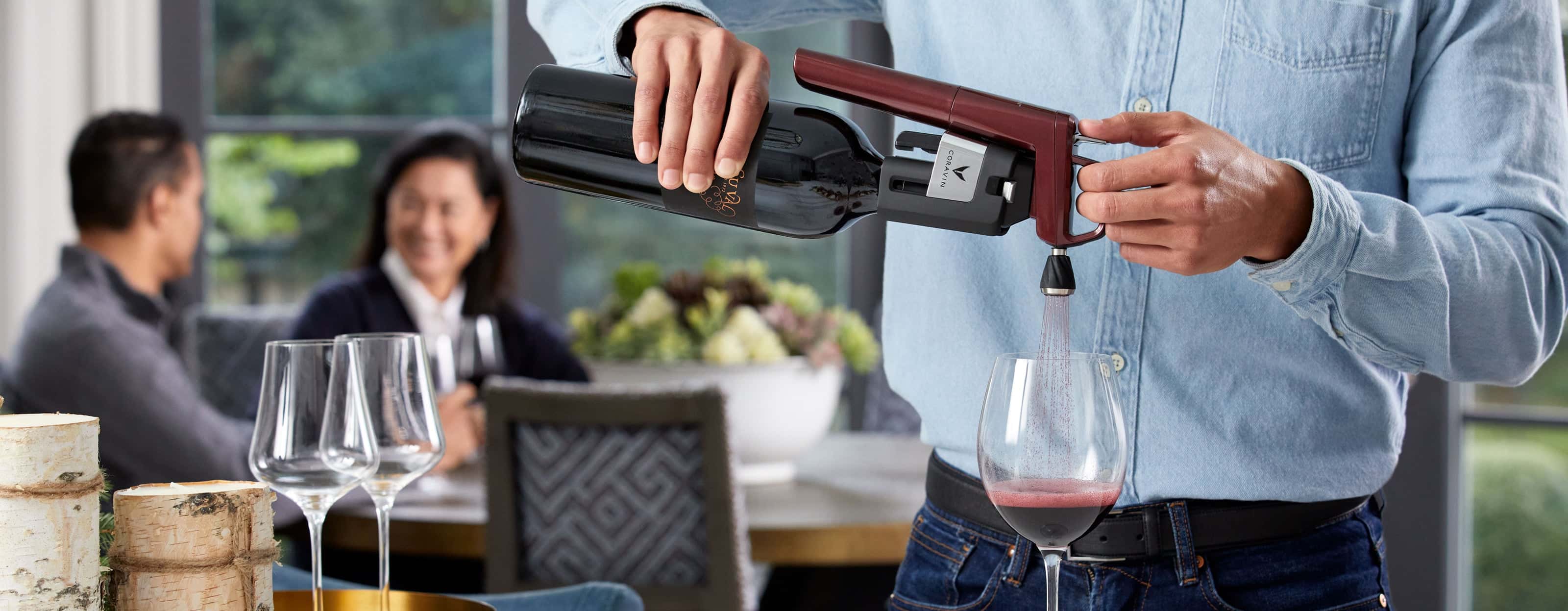 Person using Coravin Timeless Six Burgundy wine preservation system with Aerator to pour red wine, while guests enjoy conversation in the background.