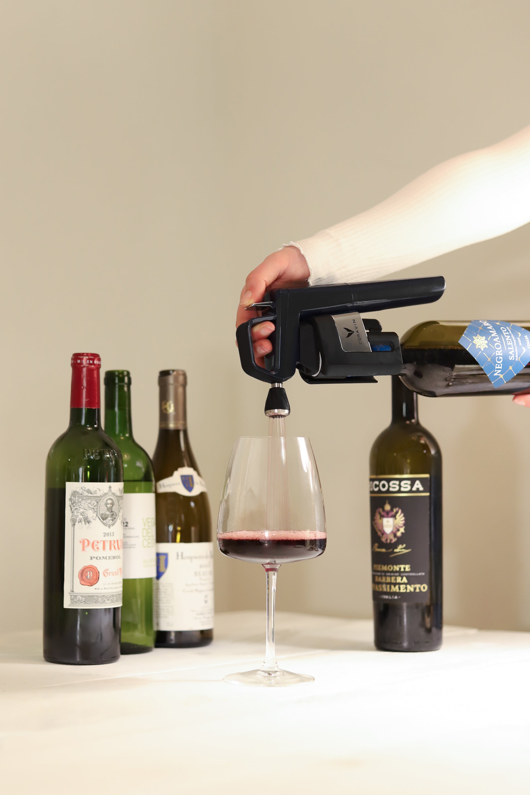Person using a Coravin Timeless Six wine preservation system to pour red wine into a glass, with four assorted wine bottles arranged on a white tablecloth in the background.