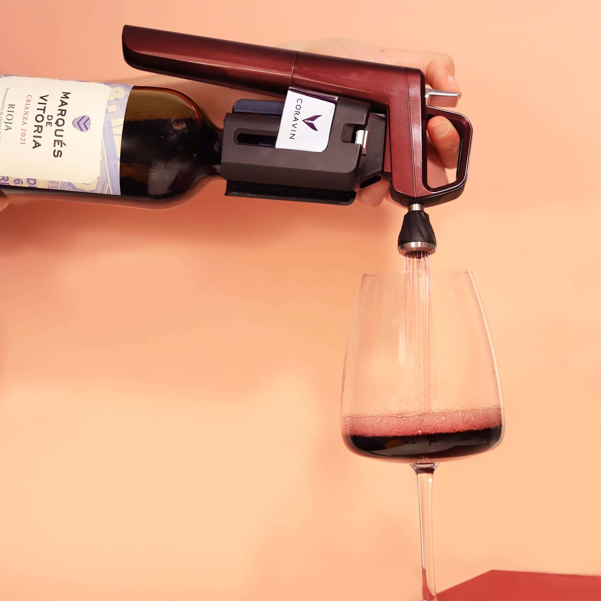 Coravin wine preservation system pouring red wine from a Marqués de Vitoria Rioja bottle into a glass against a peach-colored background.