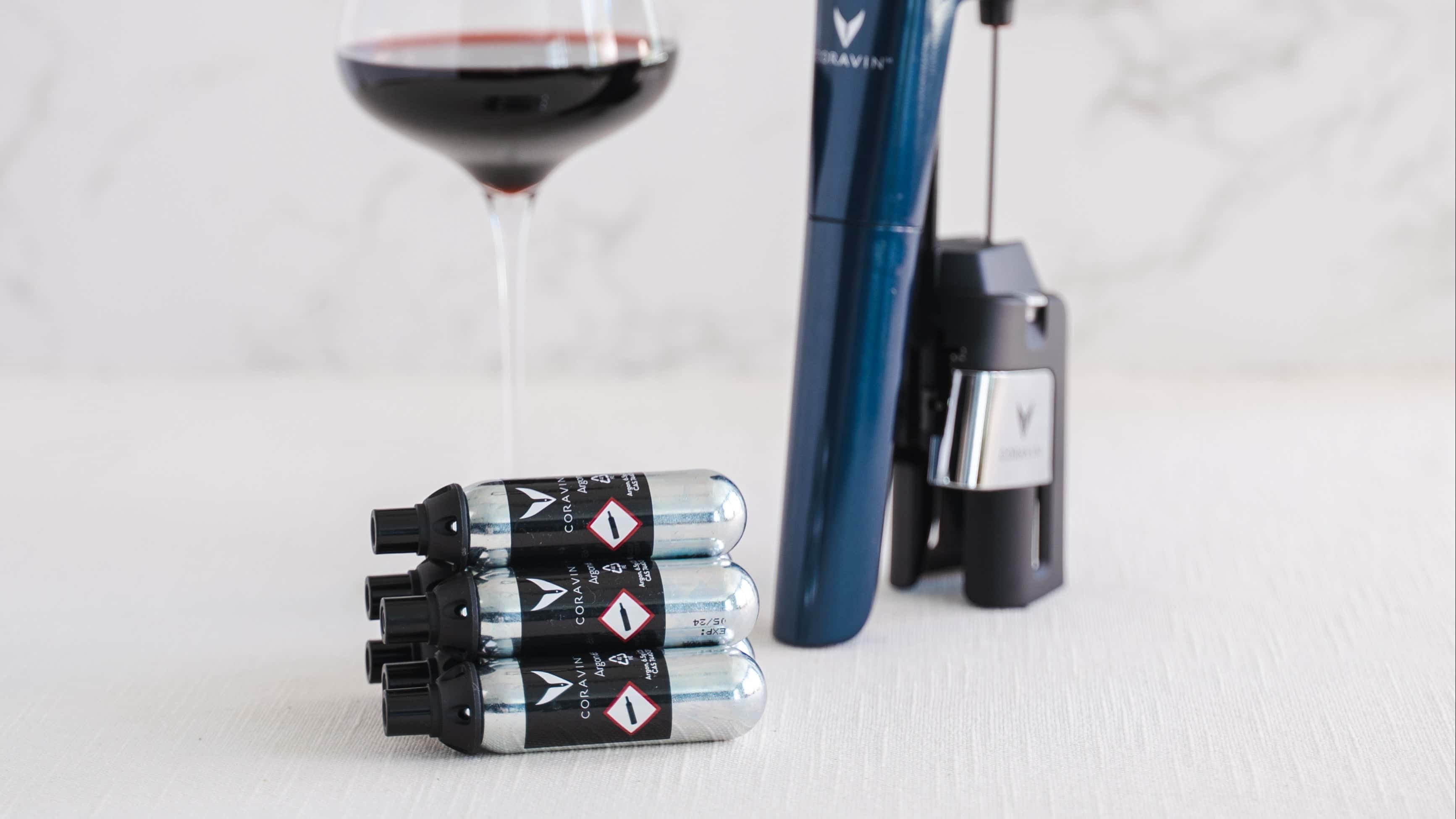 Coravin wine preservation system shown with a Timeless wine needle device, a filled glass of red wine, and a stack of argon gas capsules on a light surface.