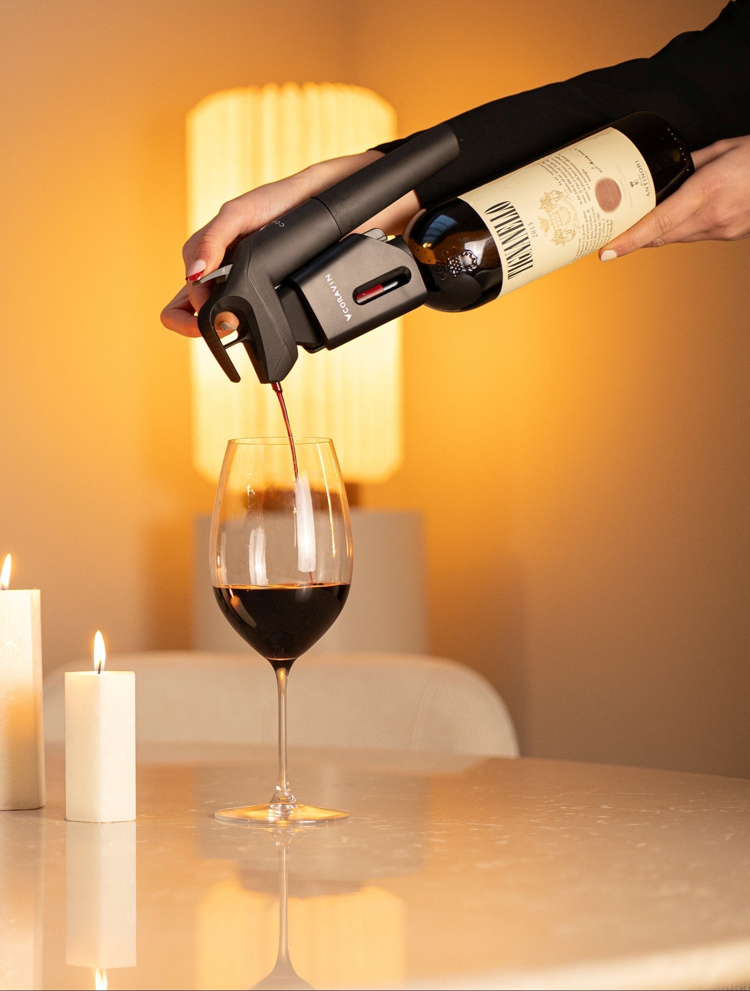 Person pouring red wine into a glass using a Coravin Timeless system, set on a marble table with lit candles and warm ambient lighting.