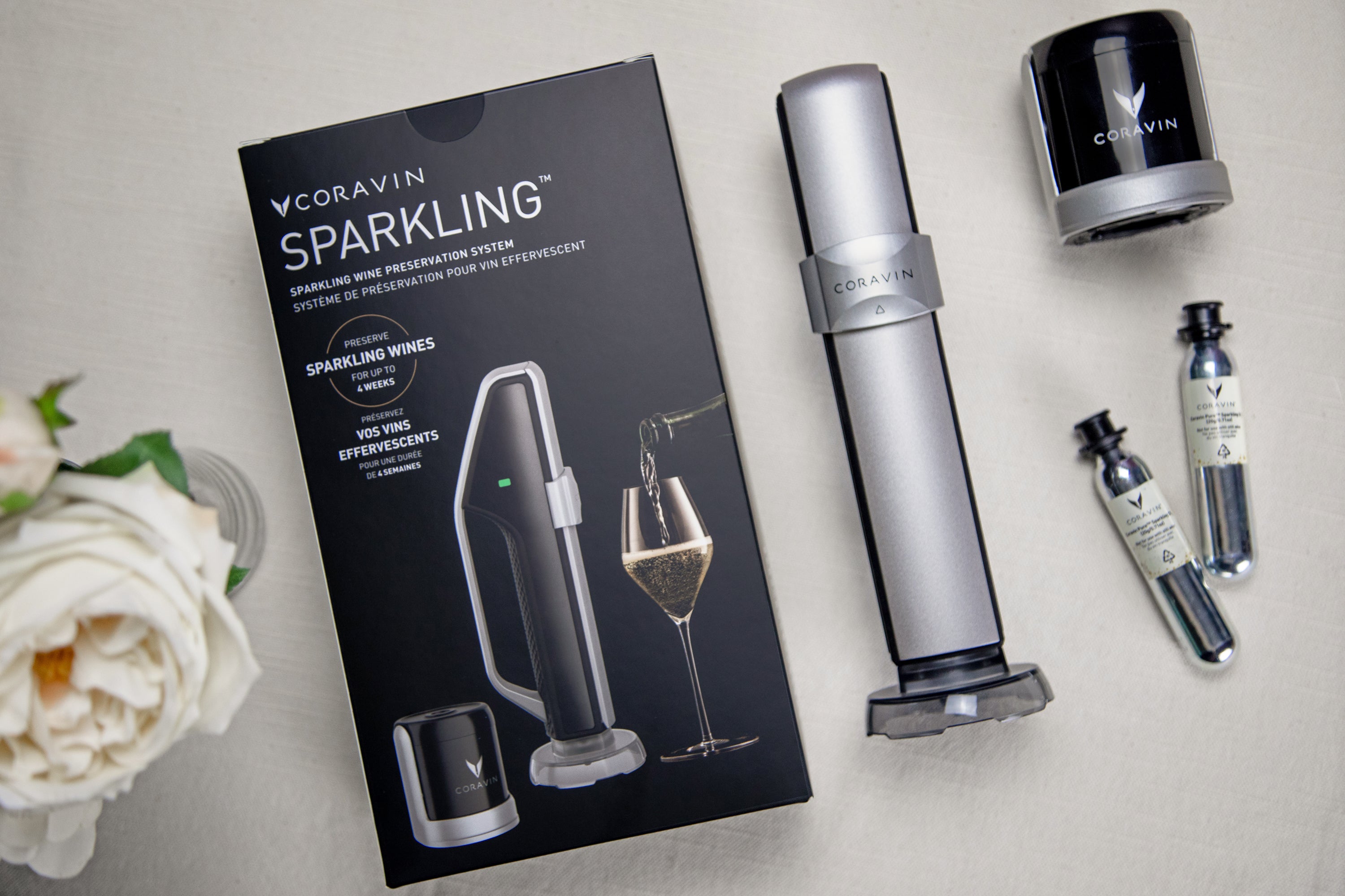 Flat lay of Coravin Sparkling Wine Preservation System packaging with device, stopper, and argon gas capsules arranged on a neutral surface next to a white rose.