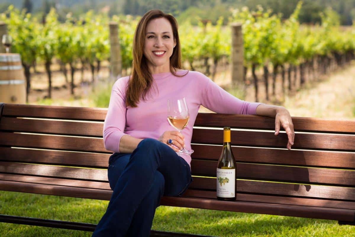 Violet Grgich sitting on a wooden bench in a vineyard holding a glass of white wine, with a bottle of Grgich Hills Estate Chardonnay beside her.