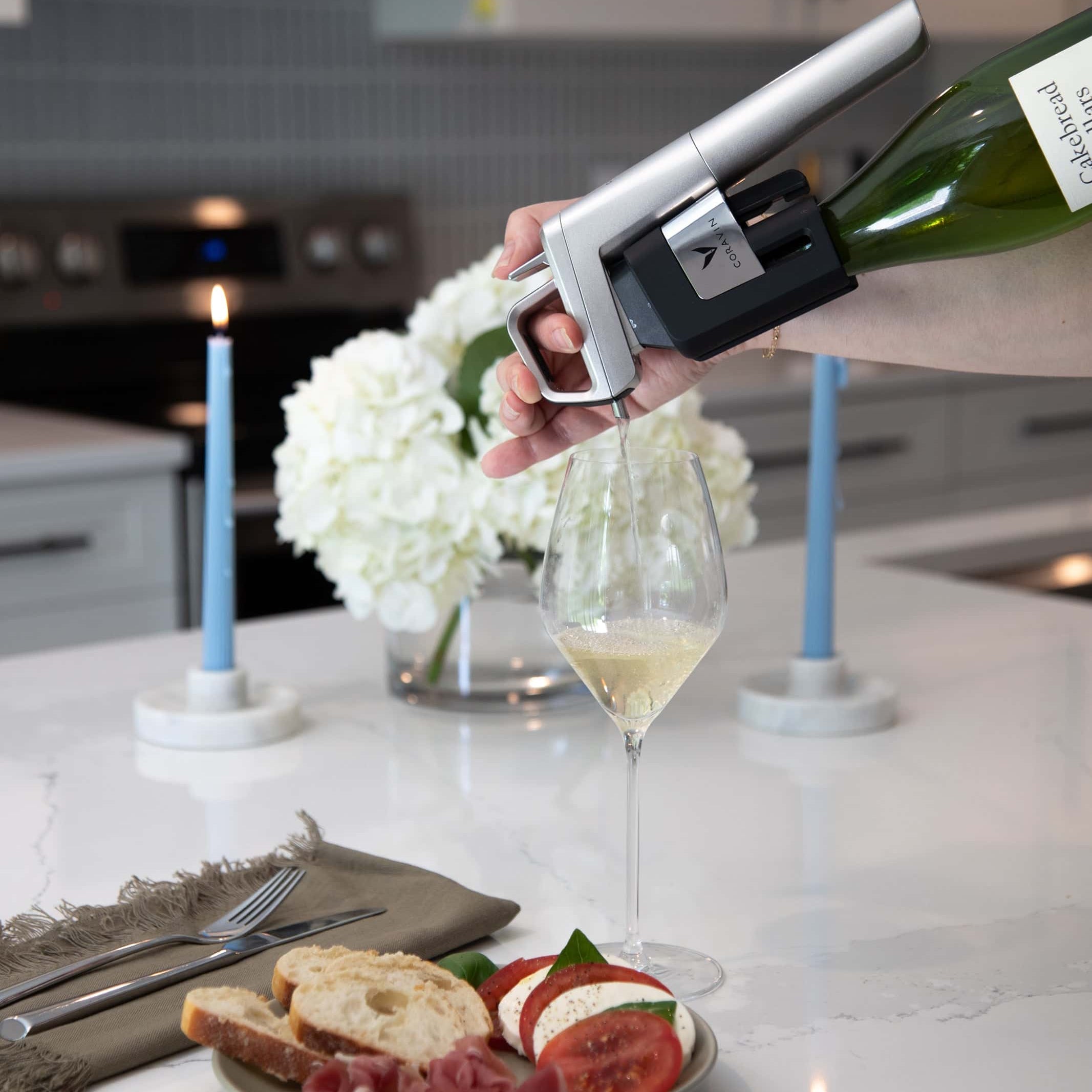 Pouring white wine using a Coravin Timeless Six preservation system into a glass on a modern kitchen counter, with a plate of prosciutto, mozzarella, tomato, and bread.