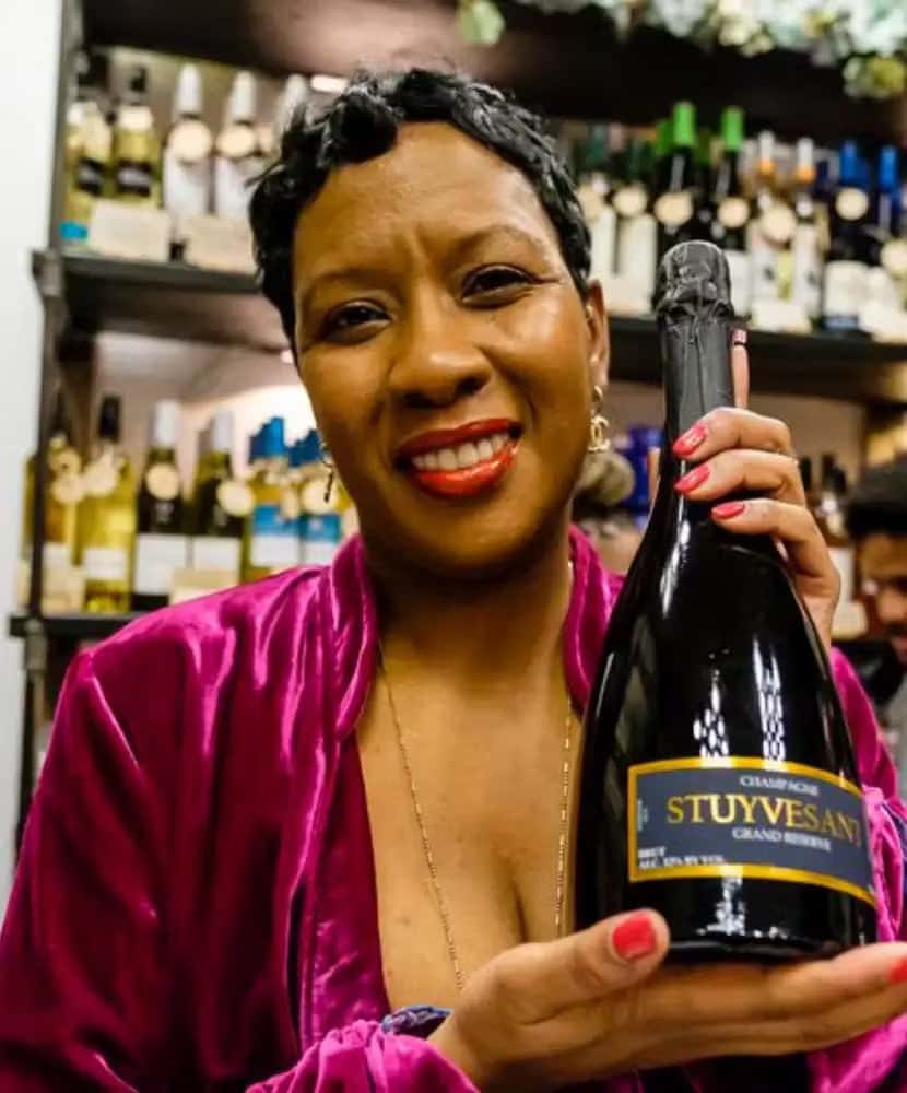 Smiling woman in a pink velvet outfit holding a bottle of Stuyvesant Champagne in a wine shop, with shelves of assorted wine bottles in the background.