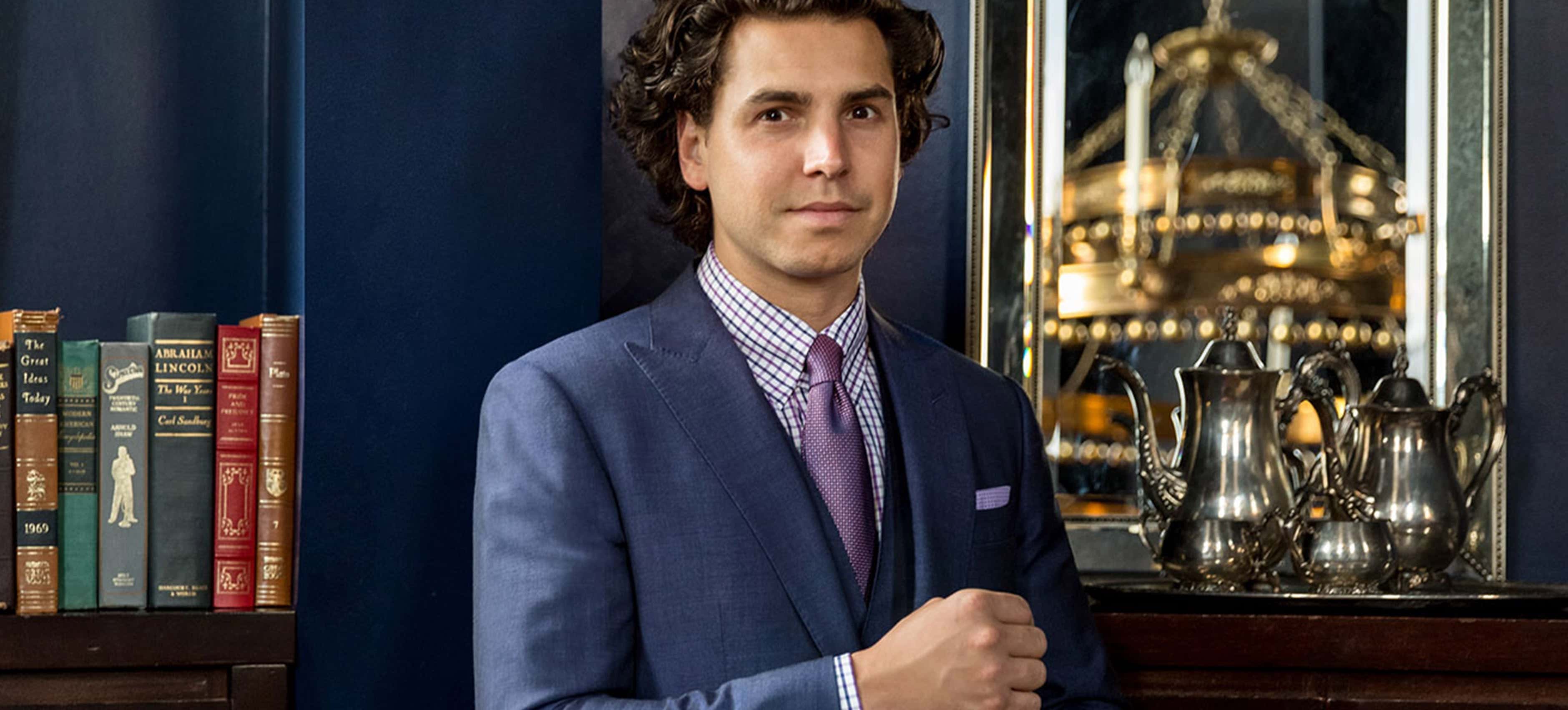 Man in a tailored blue suit with a pink tie standing beside a bookshelf and silver tea set, with a chandelier reflected in the mirror behind him.
