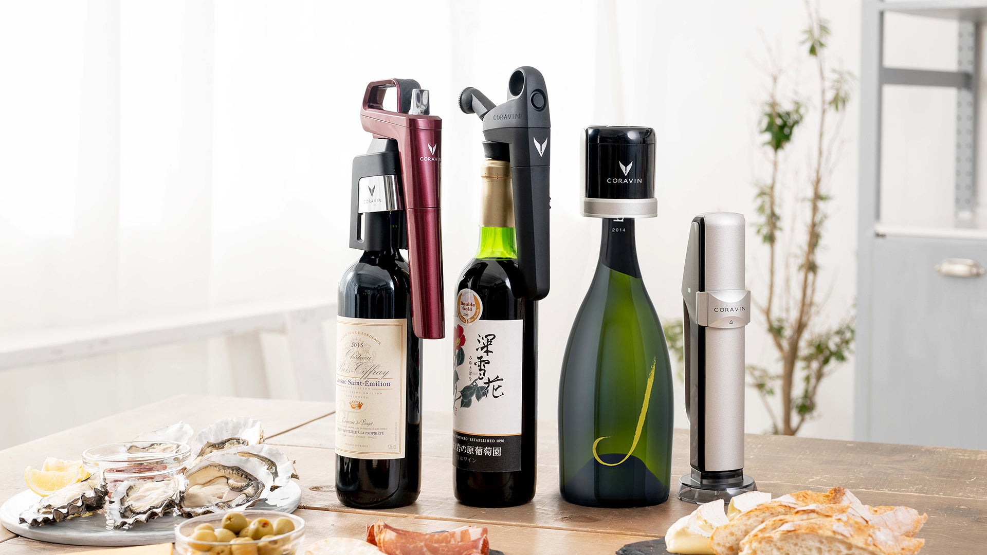 Four wine bottles with Coravin wine tools, alongside oysters and bread, set on a wooden table in a bright room.