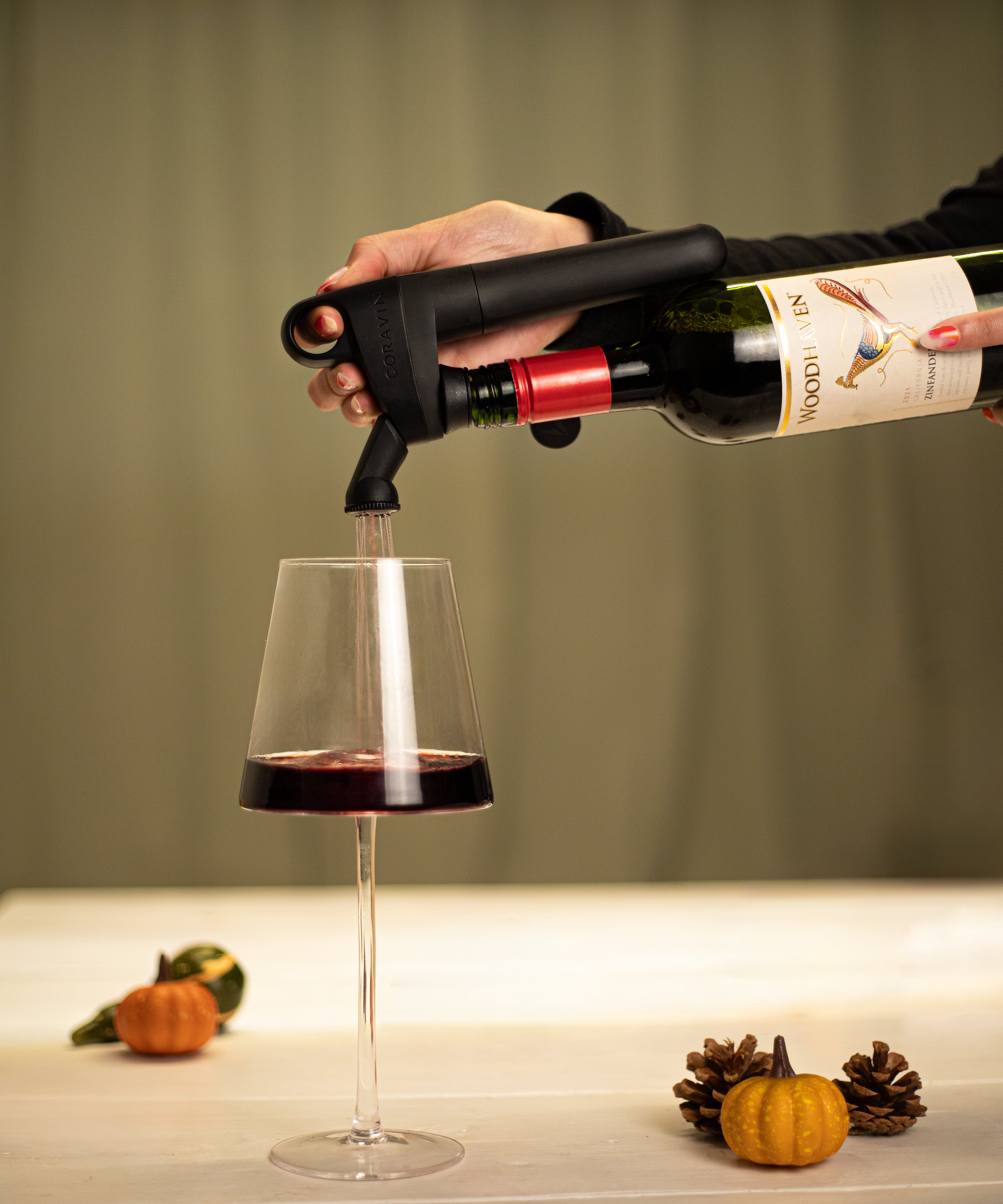 A hand uses a Coravin wine opener to pour red wine into a glass, with pumpkins and pine cones on the table.
