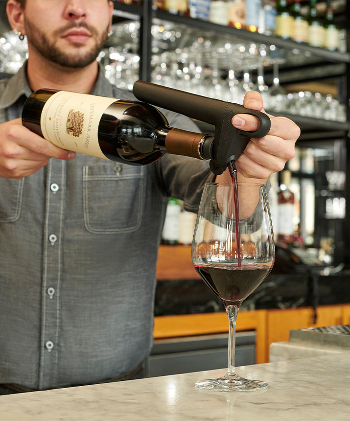 Bartender pouring red wine from a bottle using a pourer into a large glass on a marble countertop.