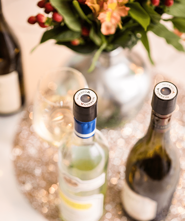 Close-up of wine bottles with Coravin closures, a glass, and a floral arrangement on a sparkling table surface.