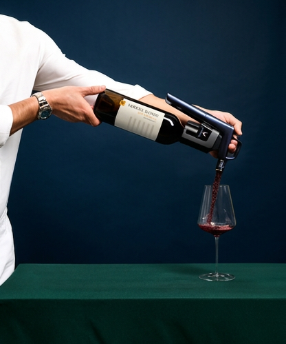 A person in a white shirt uses a Coravin to pour red wine from a bottle into a wine glass, set against a dark blue background and a green tablecloth.